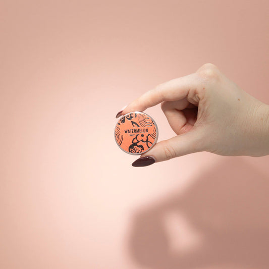 Hand holding a round, orange product with black text and graphics against a pink background