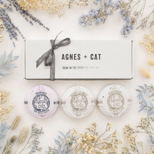 Three bath bombs with 'Agnes + Cat' branding on a decorative background with flowers and a gift box.