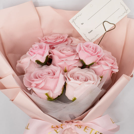 Bouquet of pink roses in a pink wrap with a card attached, on a light gray background.