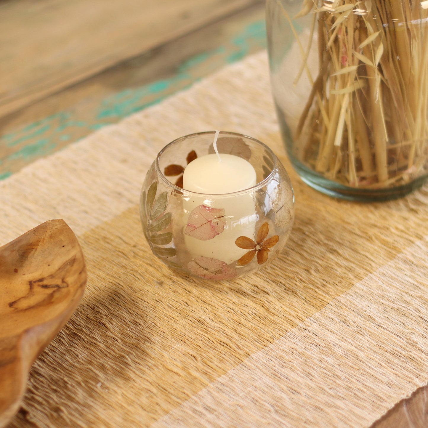 Candle in a glass holder on a woven mat with a wooden spoon and jar in the background.