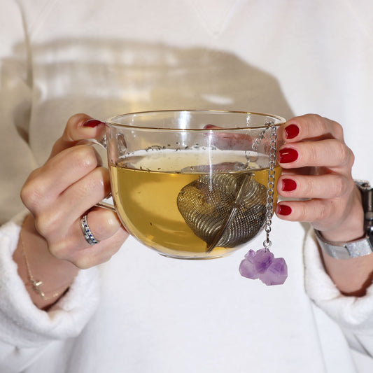 Person holding a glass mug of tea with a decorative teabag holder.