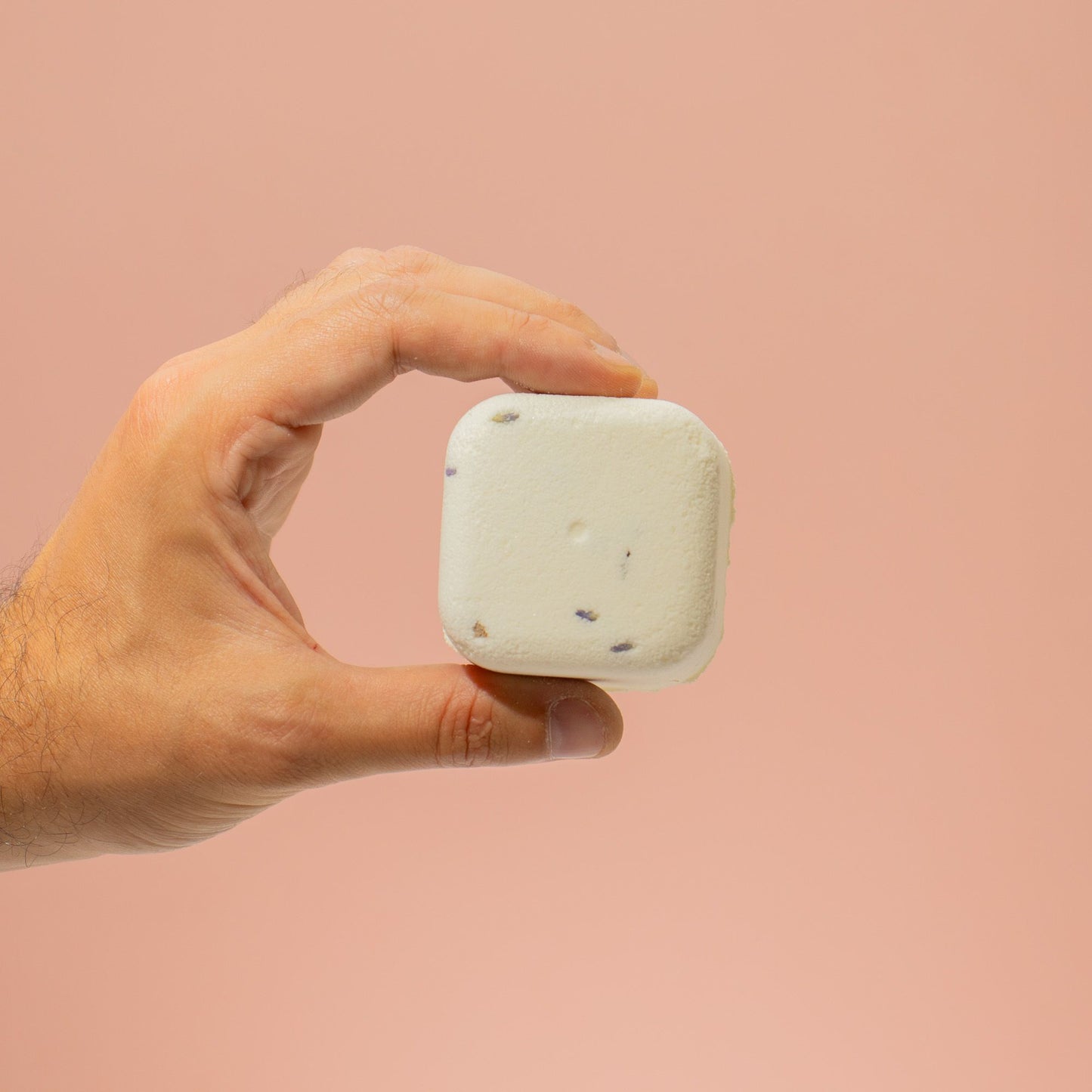 Hand holding a white square shower steamer of soap against a pink background