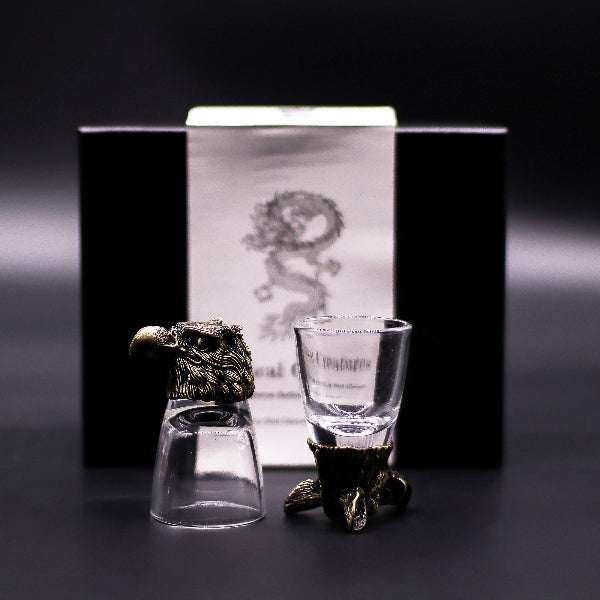 Two shot glasses with decorative stands in front of a branded box on a dark background