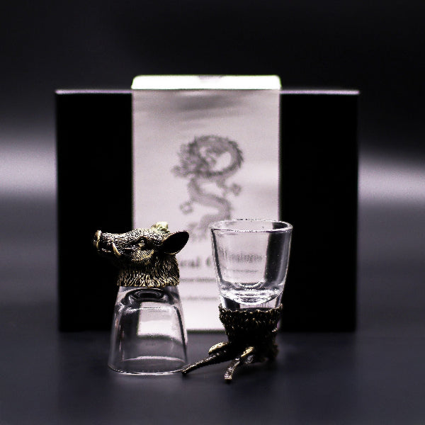 Two glass shot glasses with animal-shaped holders in front of a branded box on a dark background