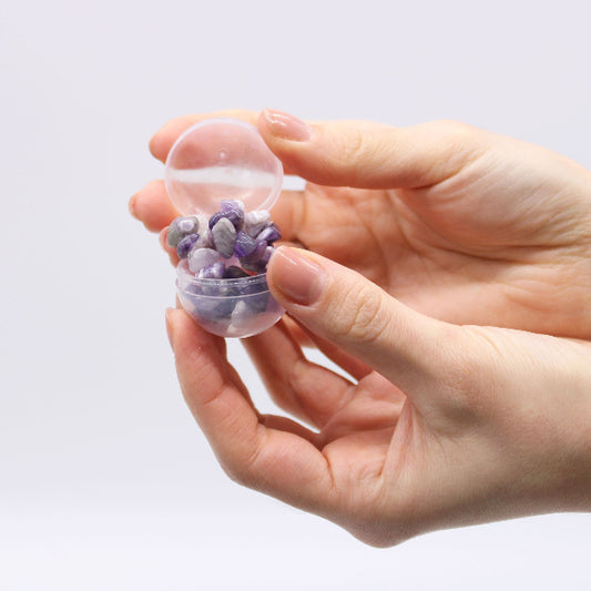 Hand holding a small transparent container filled with purple stones against a light gray background
