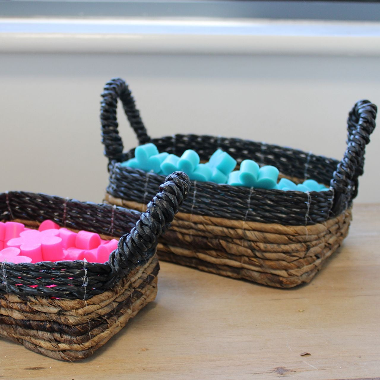 Two woven baskets with handles, one containing pink items and the other blue, on a wooden surface.