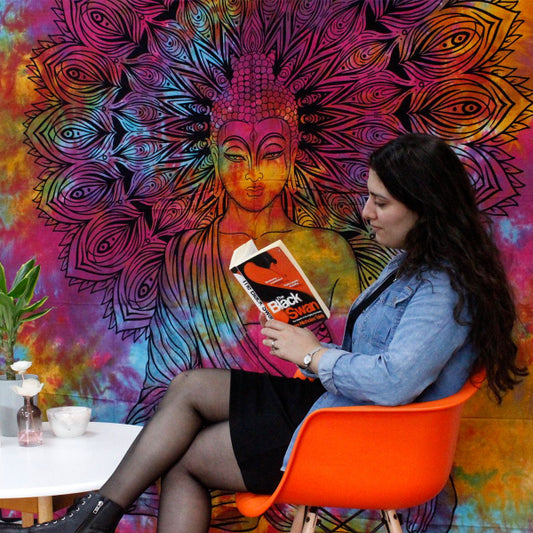 Woman sitting in an orange chair reading a book against a colorful tapestry with a Buddha design.