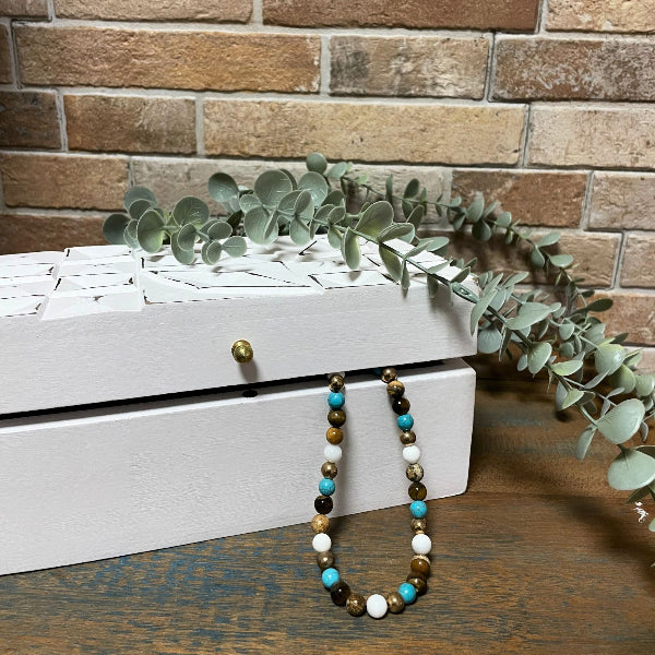 White jewelry box with a necklace and greenery against a brick wall.