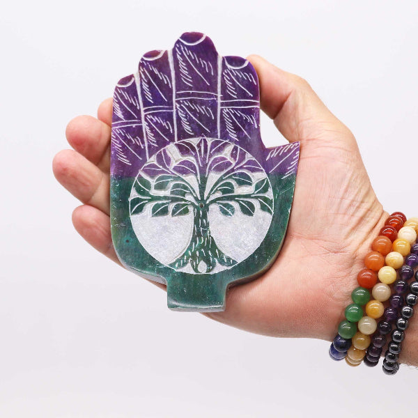 Hand holding a purple and green stone with tree of life design against a white background