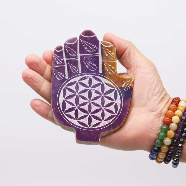 Hand holding a purple crystal with a flower of life design against a white background