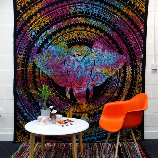 Colorful tapestry with an elephant design hanging on a wall above a table and chair.