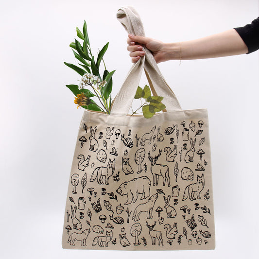 Beige tote bag with black animal illustrations held by a hand against a white background