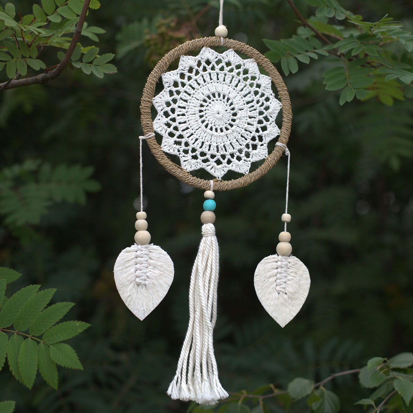 Decorative dreamcatcher with white crochet design, leaves, and tassel against a green leafy background