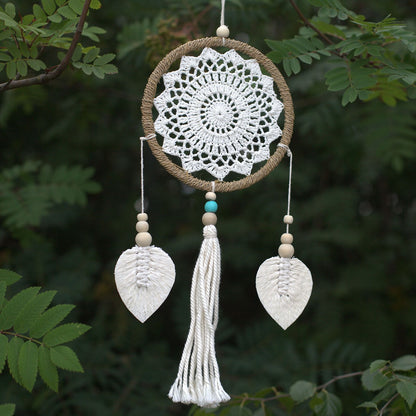 Decorative dreamcatcher with white crochet design, leaves, and tassel against a green leafy background
