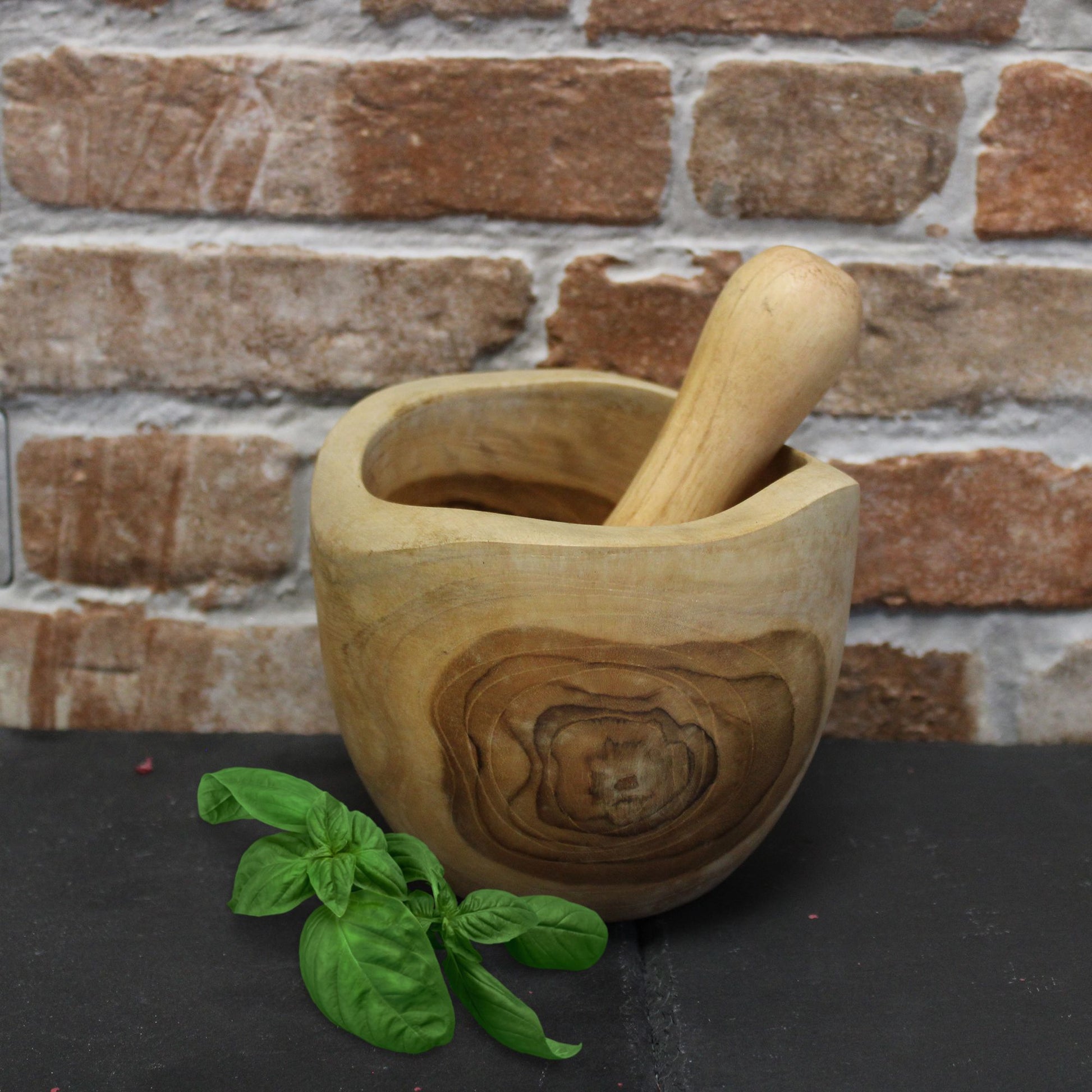 Wooden mortar and pestle with basil leaves on a dark surface against a brick wall.