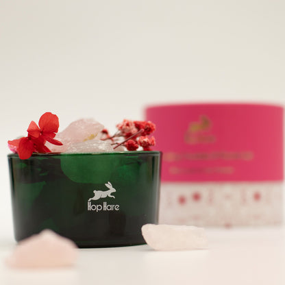 Green container with flowers and crystals on a white surface, with a blurred pink box in the background.