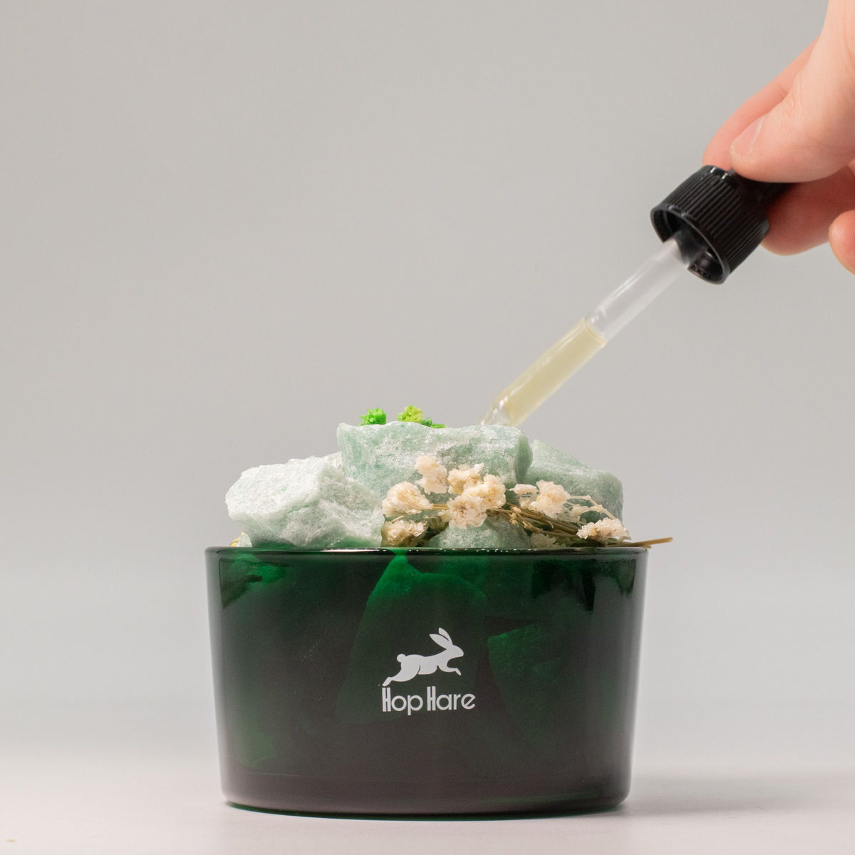 Hand using a dropper to add liquid to a green container with a brand logo on a gray background