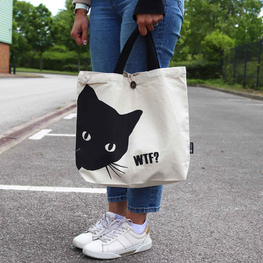Person holding a tote bag with a black cat design and 'WTF?' text on a street.