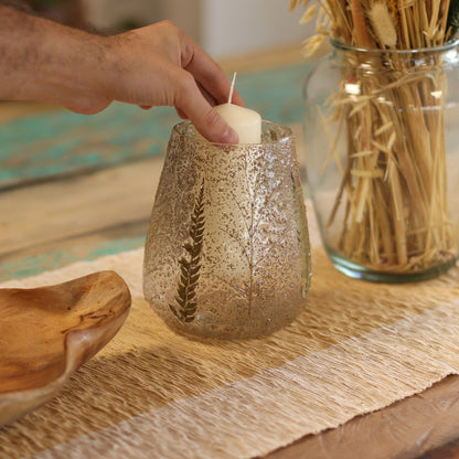 Hand lighting a candle in a textured glass holder on a table with a wooden bowl and vase in the background.