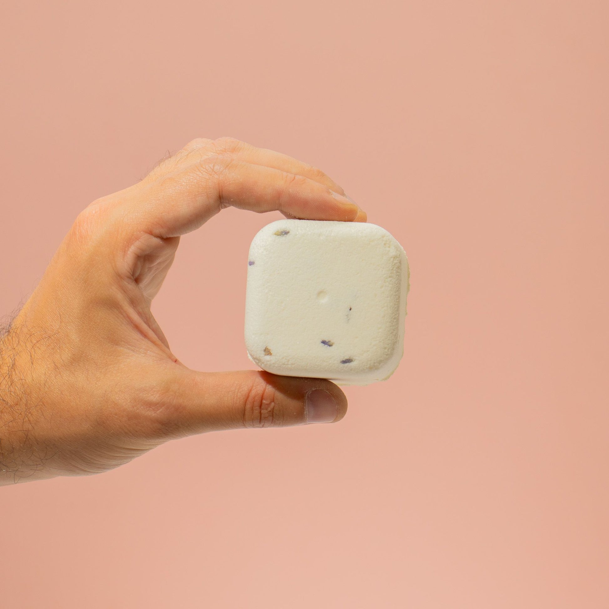 Hand holding a white square shower steamer of soap against a pink background