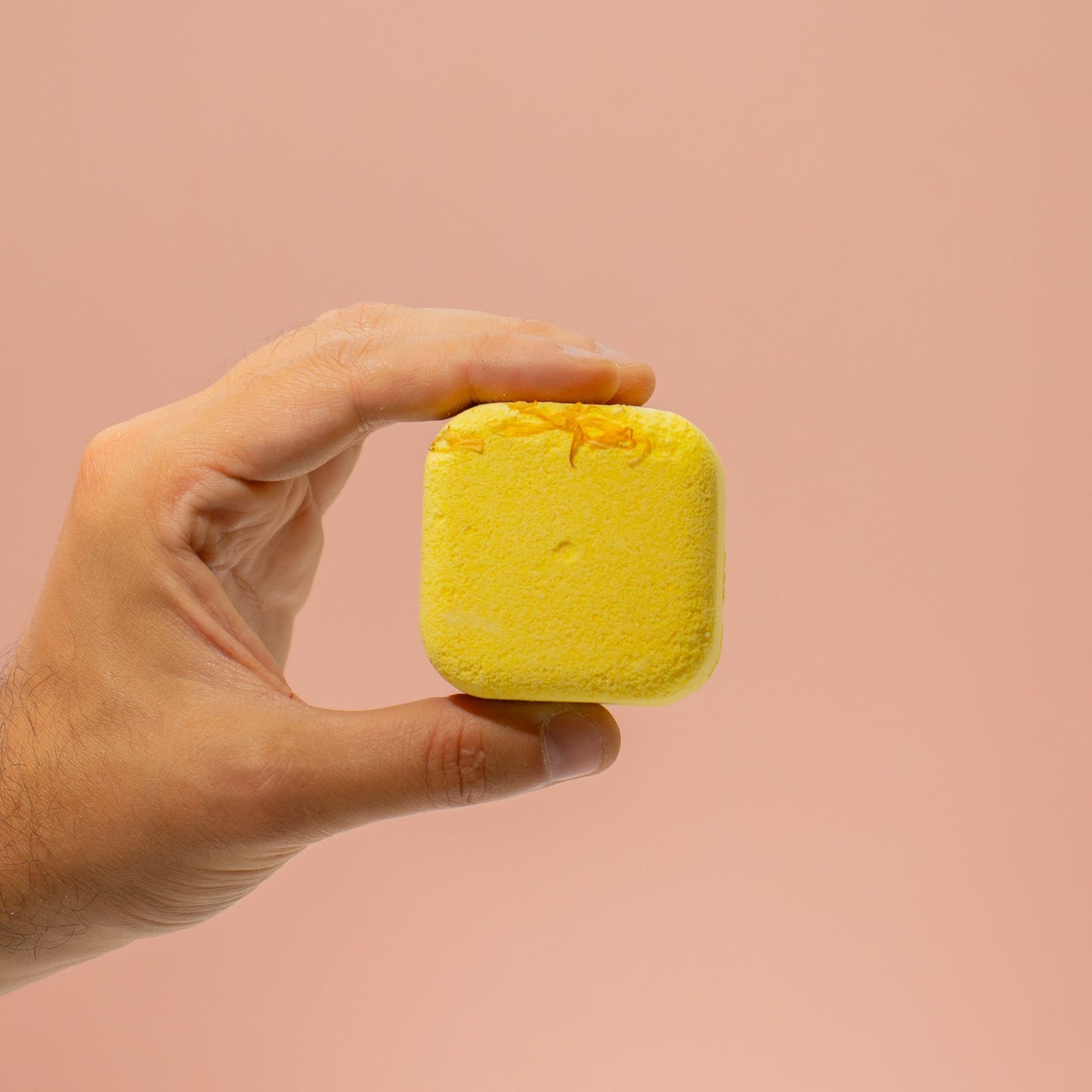 Hand holding a yellow square bath bomb against a pink background