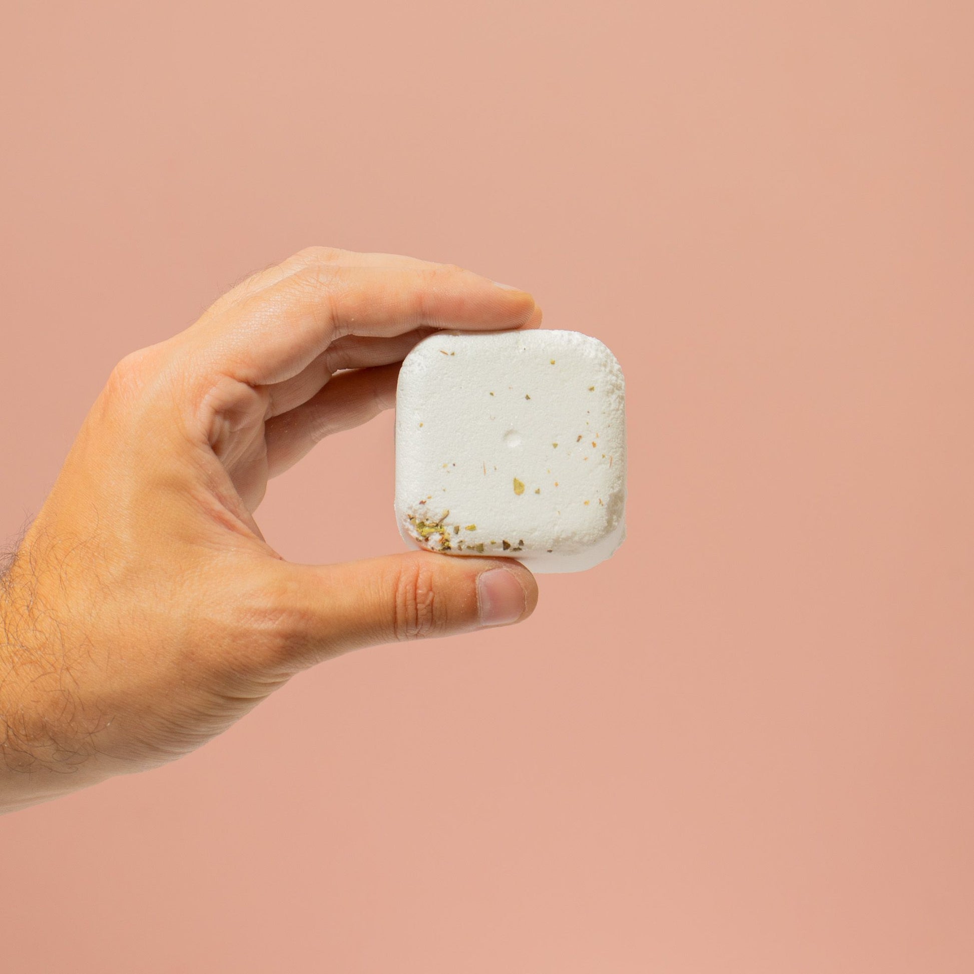 Hand holding a square, white bath bomb with gold specks against a pink background