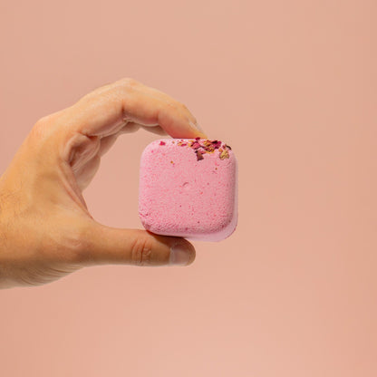 Hand holding a pink bath bomb with rose petals against a beige background