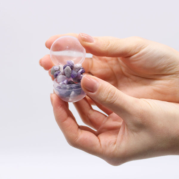 Hand holding a small transparent container filled with purple stones against a light gray background