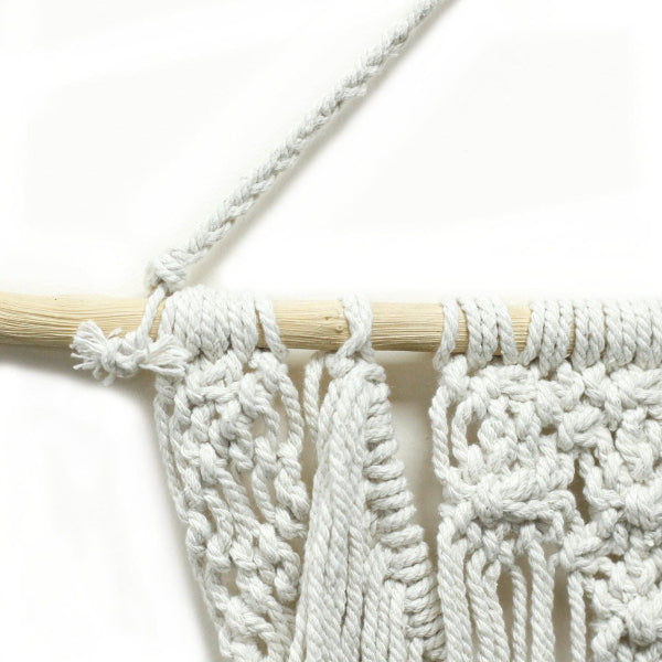 Close-up of a white macrame wall hanging with wooden dowel on a light background