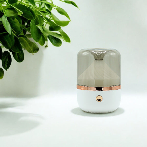 Small cylindrical humidifier with rose gold accents on a white surface with a plant in the corner.
