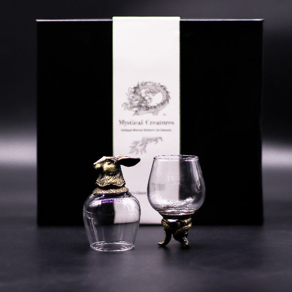 Two glass decanters with gold animal-shaped stoppers in front of a black box labeled 'Mystical Creatures'.