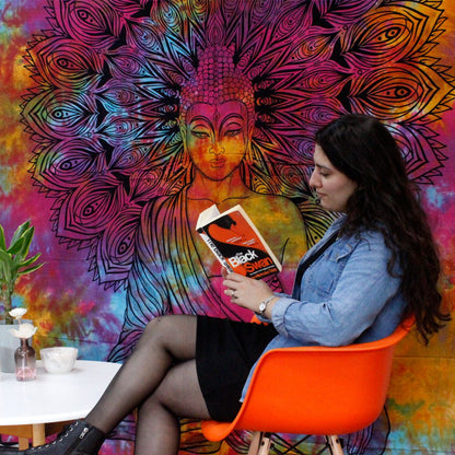 Woman sitting in an orange chair reading a book against a colorful tapestry with a Buddha design.