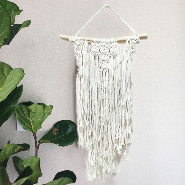 White macrame wall hanging on a light gray wall with green leaves.