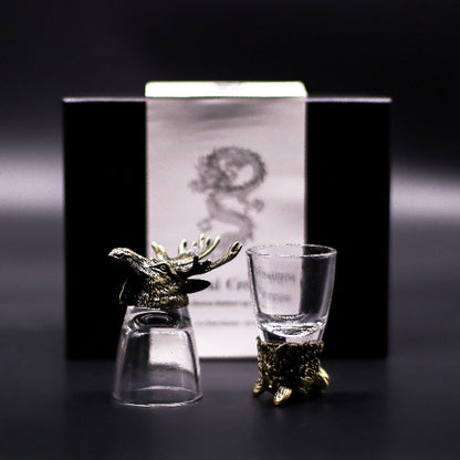 Two glass shot glasses with gold deer heads on a dark background