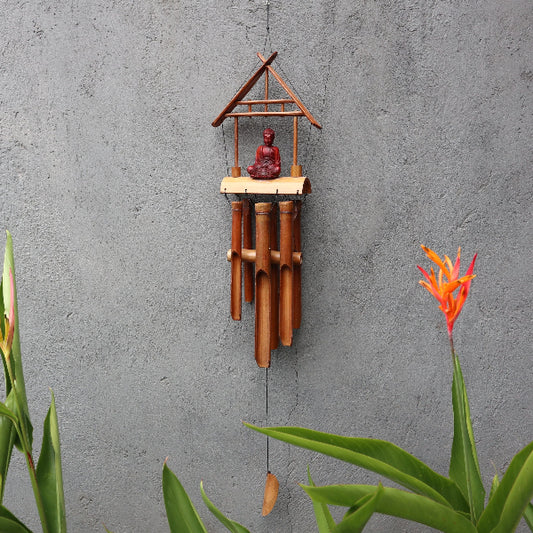 Bamboo wind chime with a small statue on a gray wall with plants.