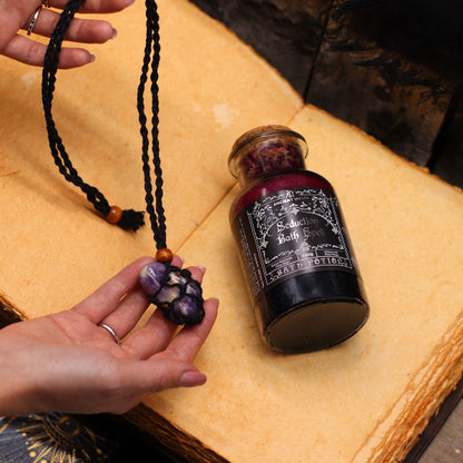 Hand holding a necklace with a small jar labeled 'Seduction Bath Salts' on a textured surface.