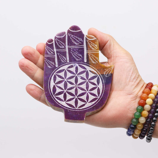 Hand holding a purple crystal with a flower of life design against a white background