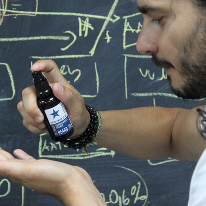 Person holding a bottle of beard oil in front of a chalkboard with drawings.